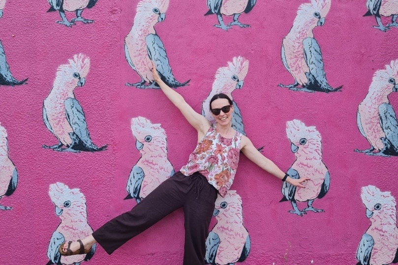 A woman in a pink floral top and black pants standing in front of a pink mural depicting multiple birds