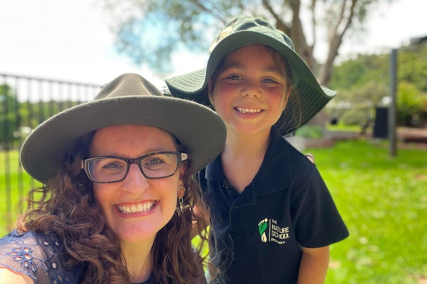 A female teacher smiling outdoors, wearing a wide brimmed hat, with a young female student beside her.