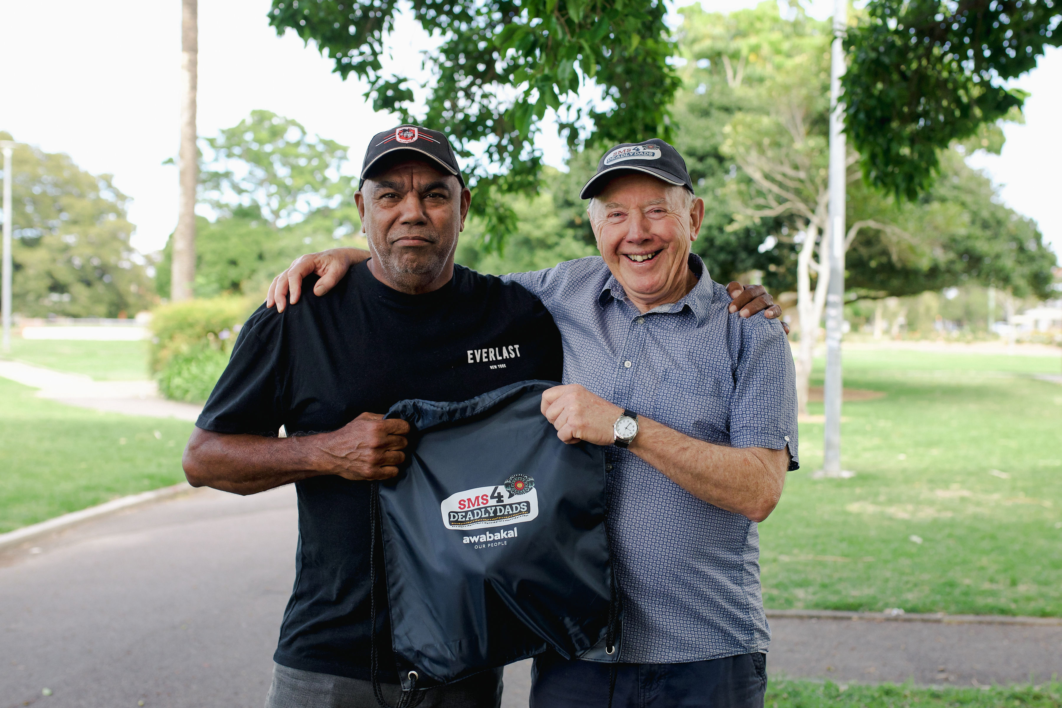 Two men, one Indigenous the other non-Indigenous, stand together and hold up a navy bag.