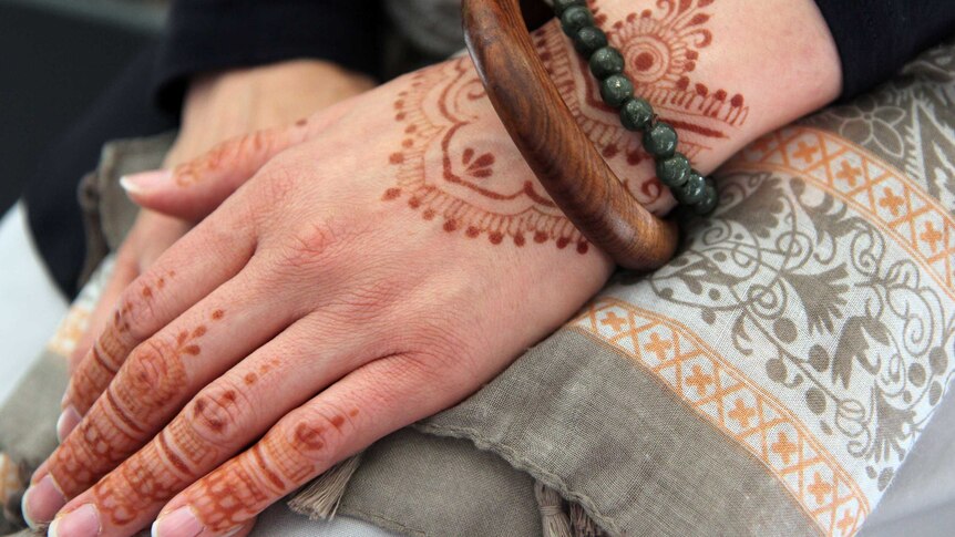 A woman's folded hands. One hand features a wooden bangle and green beads and  is painted with henna in an intricate pattern.