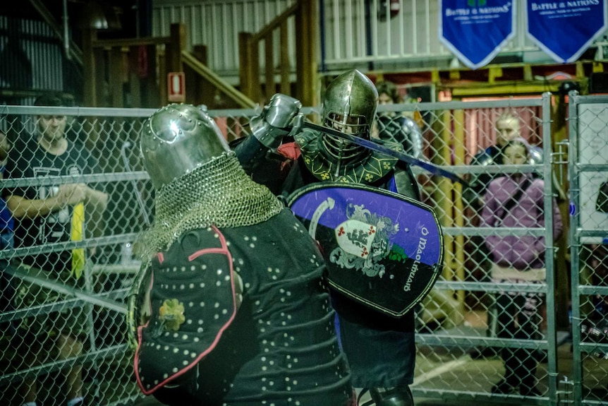 Two fighters in armour and shields fight in a caged arena