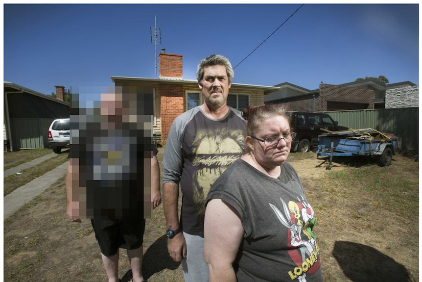 Two men, one of whom is blurred, and a woman stand in front of a house.