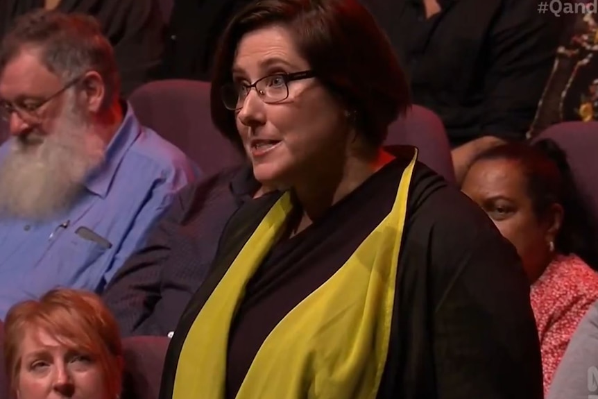 A woman wearing glasses and a yellow scarf speaks.