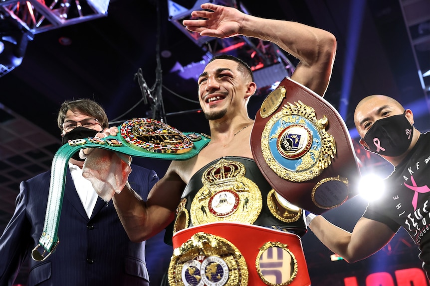 Teofimo Lopez holds four boxing championship belts and waves, smiling
