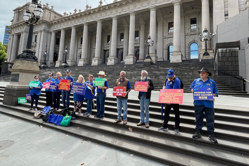 Eleven people stand on the steps outside Melbourne's Parliament House holding protest signs