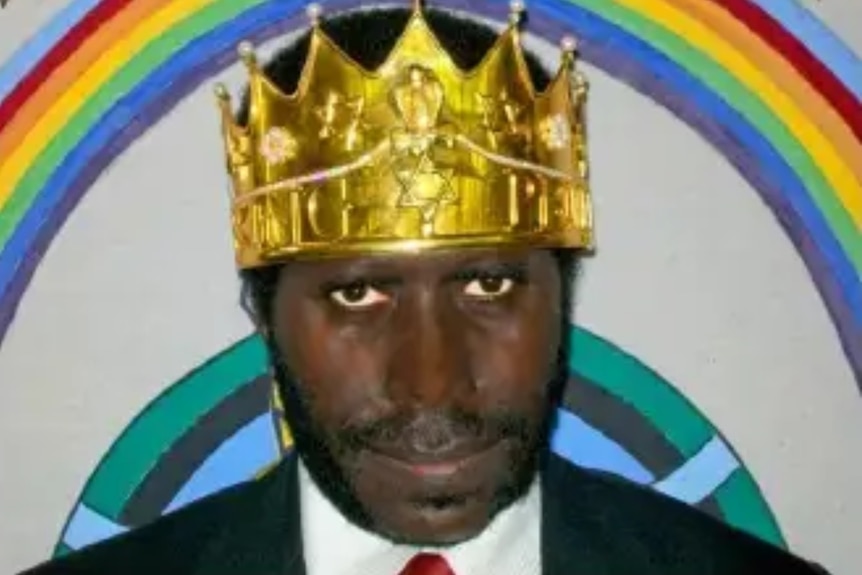 Noah Musingku is shown wearing a fake crown, standing in a suit in front of a painted rainbow.
