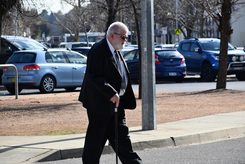A white-haired man in a suit walks across a road, assisted by a black cane.