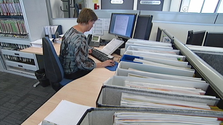 Video still: Public servant working behind a desk in an office in Canberra - generic no faces - vertical files in foreground.
