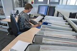 Video still: Public servant working behind a desk in an office in Canberra - generic no faces - vertical files in foreground.