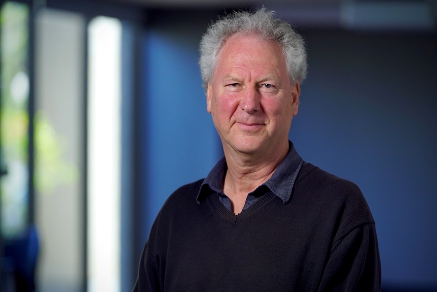 A man with white hair and a dark coloured shirt and jumper stares at the camera