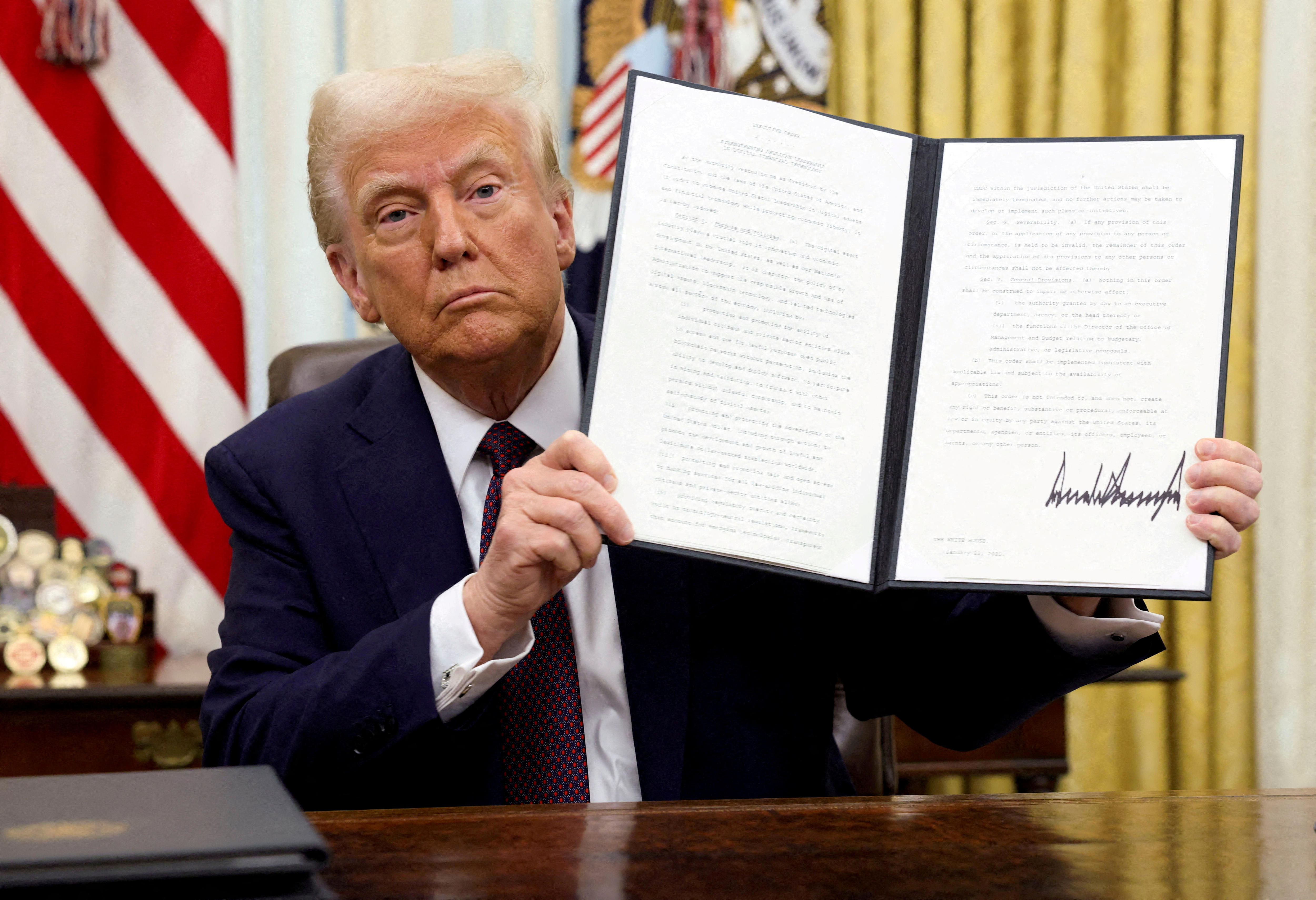 Donald Trump in a dark suit jacket sitting at a wooden desk while holding up a signed executive order in a binder