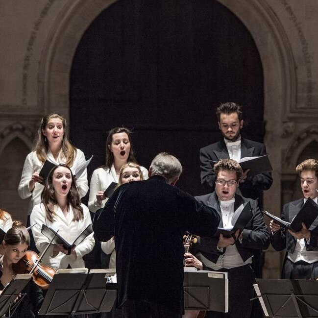A photograph of choristers performing with an early music ensemble in a cathedral-like space, with a conductor out the front.