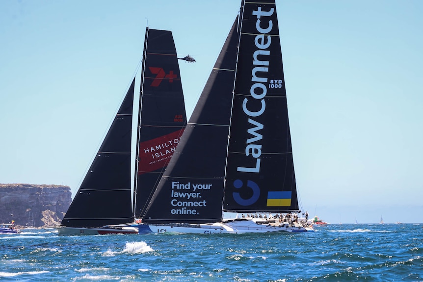 Two big boats move through the water towards the Sydney Heads with sails up and a helicopter in the background.  