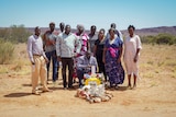 An African family of around 10 people stands around a memorial among a desert landscape