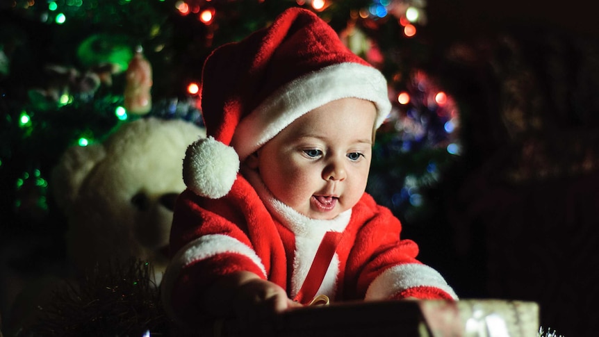 A baby in a Santa outfit looks expectantly at presents in front of a Christmas tree