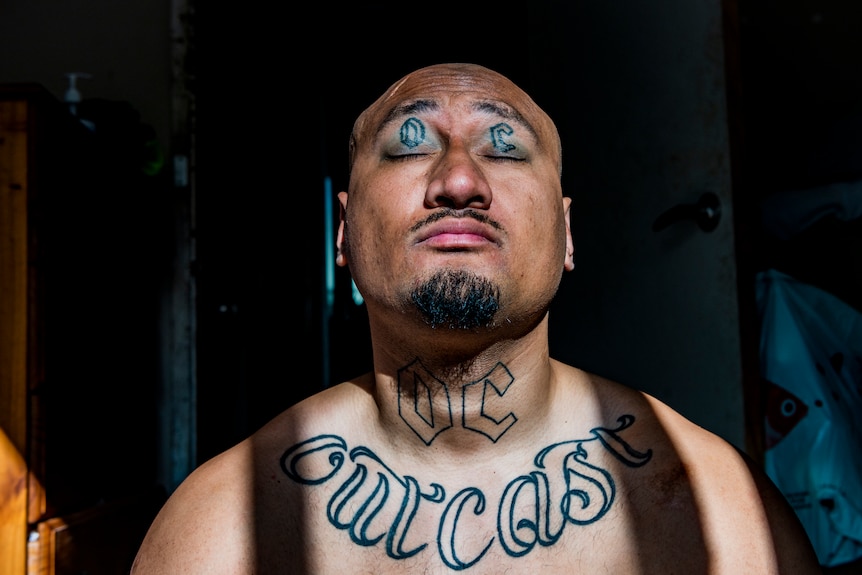A man closes his eyes revealing tattoos of the letters "OC" on his eyelids. He has the word "Outcast" tattooed on his chest.