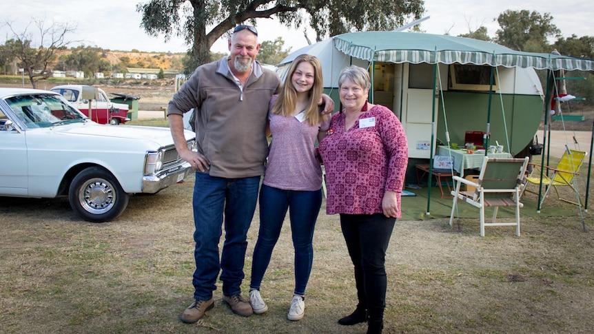 A man, woman and teenage girl stand in front of a vintage green caravan and classic car at a caravan park.
