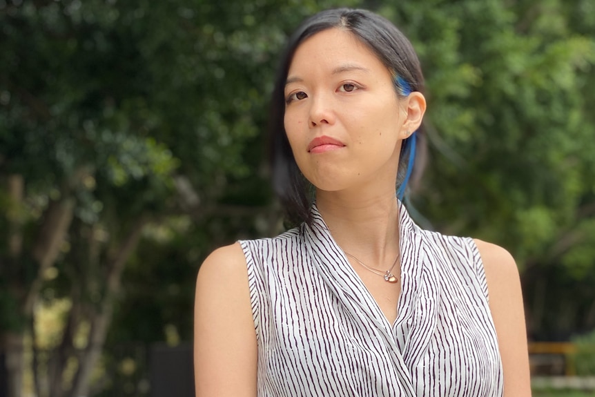 An Asian woman wearing a black and white sleeveless blouse stands in front of trees.