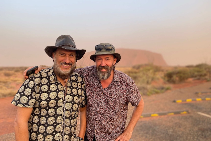 Claudio Villella and Andrew Kuhene have their arms around each other and smile for the camera. Behind them is a road and Uluru.