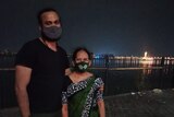 A man in a black tshirt and mask stands with his arm around an older woman in a sari and mask. A city and river in the backgroun