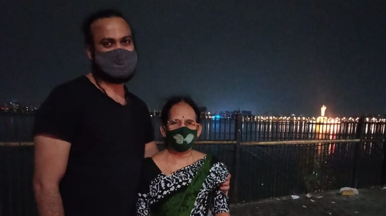 A man in a black tshirt and mask stands with his arm around an older woman in a sari and mask. A city and river in the backgroun