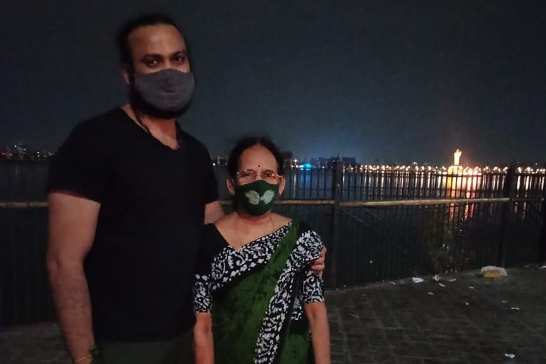 A man in a black tshirt and mask stands with his arm around an older woman in a sari and mask. A city and river in the backgroun