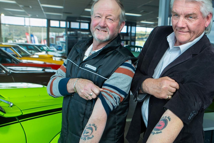Two men lift sleeves to reveal matching tattoos