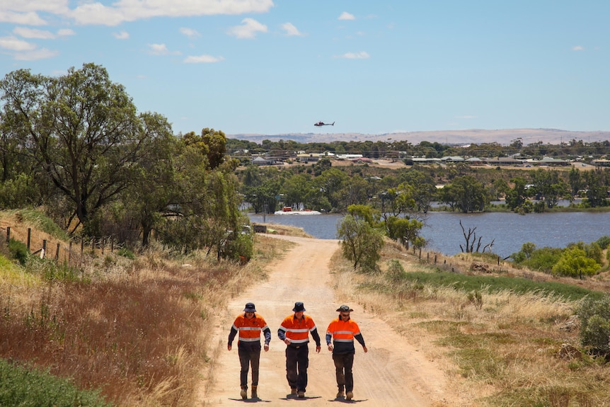 Three people in orange and white high-vis tops walk along a dirt path. In the distant background is a helicopter over a river