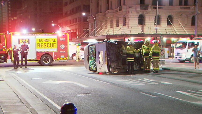 A van full of e-scooters sits on its side in the middle of the road as firemen assess the scene
