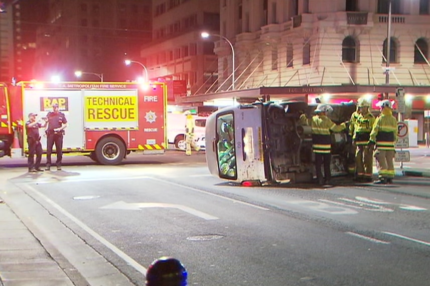 A van full of e-scooters sits on its side in the middle of the road as firemen assess the scene