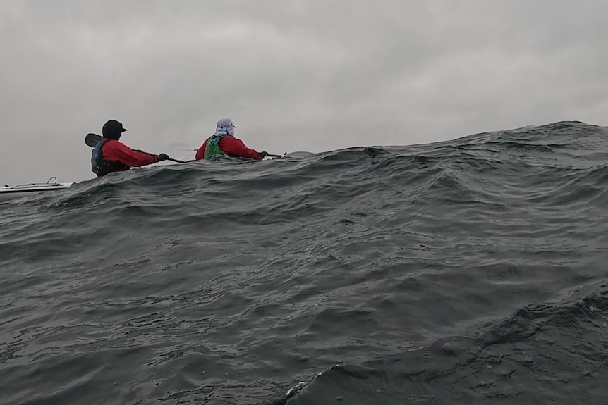 Two people wearing caps, life jackets kayak as turbulant water rises, overcast, mist .
