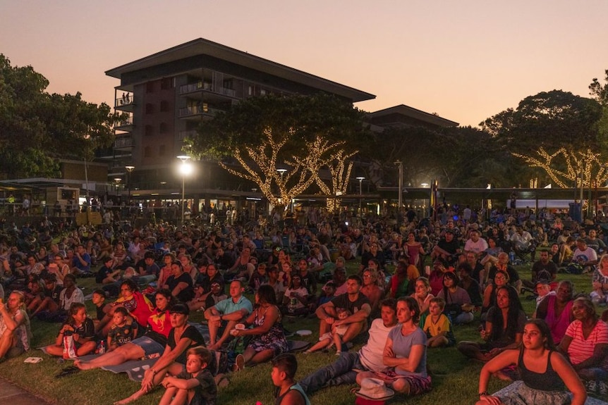 People sit on the grass at an outdoor concert at sunset.