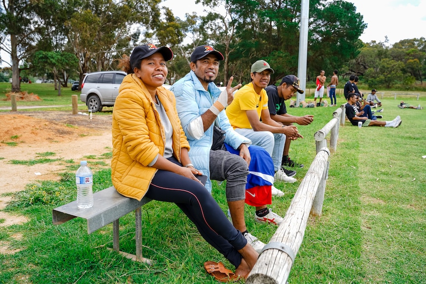 Smiling people sit on a bench on the edge of a sporting ground.