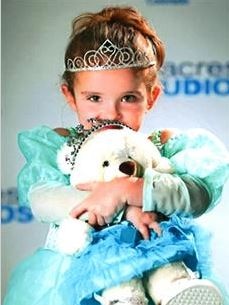 Olivia K Gant dressed in a blue dress wearing a plastic tiara and holding a teddy bear.