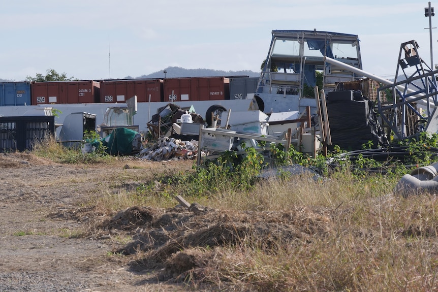 A messy yard, an old boat in a storage yard with shipping containers in the background.