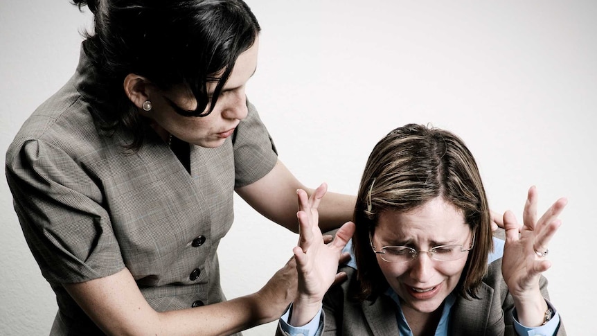 A woman leaning over another woman who is sitting at a typewriter with a frustrated look as she gestures with her hands.