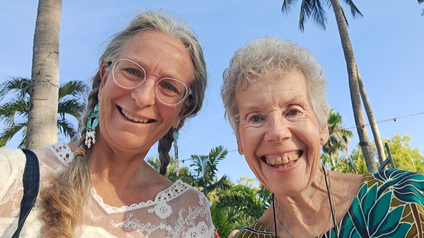 Two grey haired women smile at camera, palm trees and blue sky in background