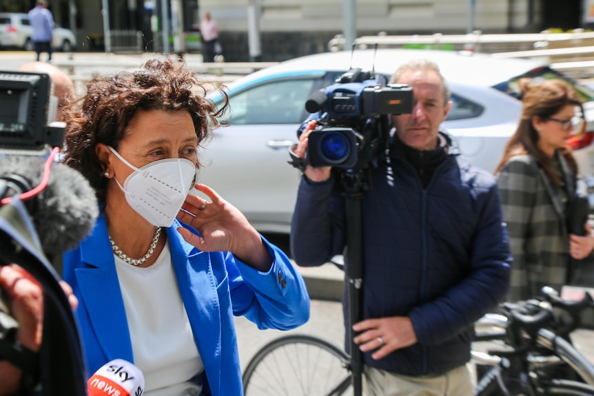 MP Monique Ryan wears a face mask and a blue suit outside court in the sunshine, surrounded by media.