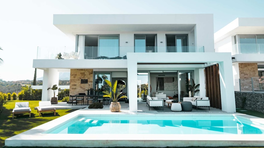 A grand modern double-storey white home with a porch overlooking an outdoor pool on a sunny day.