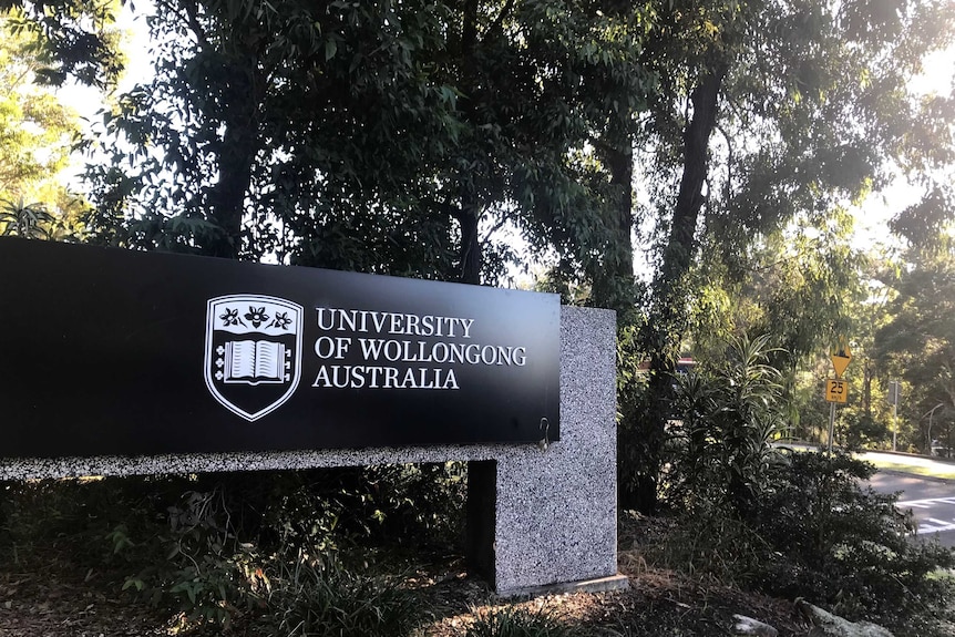 Long black entrance sign at gateway to University of Wollongong on granite stone with trees behind.