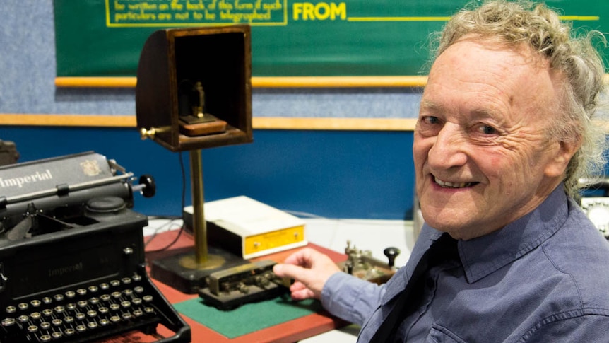 An older man sits in front of a telegraph machine and typewriter.