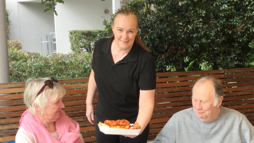 A woman with hair pulled back in a ponytail serves fish and chips to an elderly couple at an outside dining area.