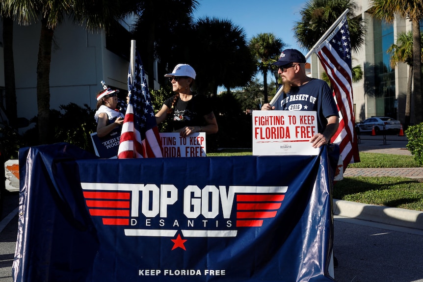 Two people dressed in Ron DeSantis merchandise stand behind a blue, red and white flag that says "TOP GOVE DESANTIS"