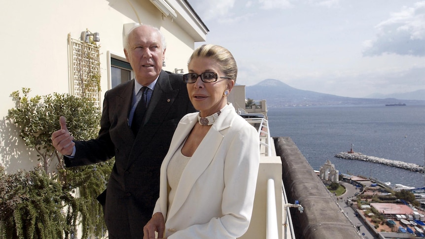 A man dressed in a suit and a woman dressed in a white suit stand on a balcony next to a scenic view of the ocean.