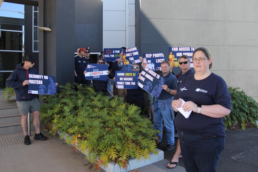 A woman in a union shirt stands in front of a group of men holding placards outside an office building.