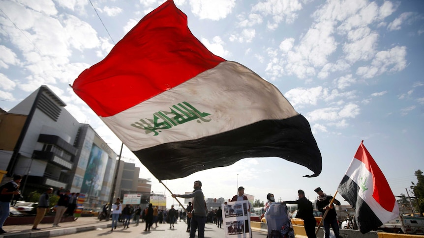 A crowd of students are pictured in the middle of a road on a cloudy day, with a large Iraqi flag flying in the centre.