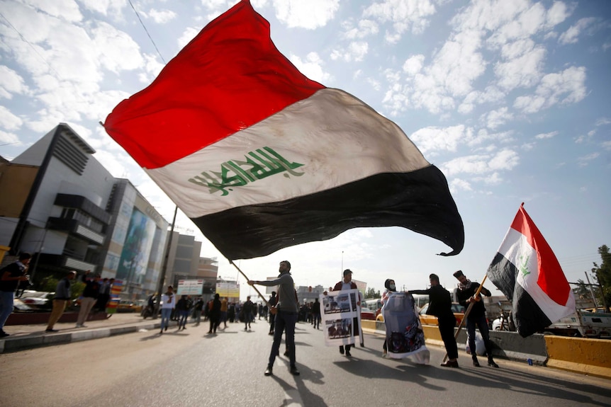 A crowd of students are pictured in the middle of a road on a cloudy day, with a large Iraqi flag flying in the centre.