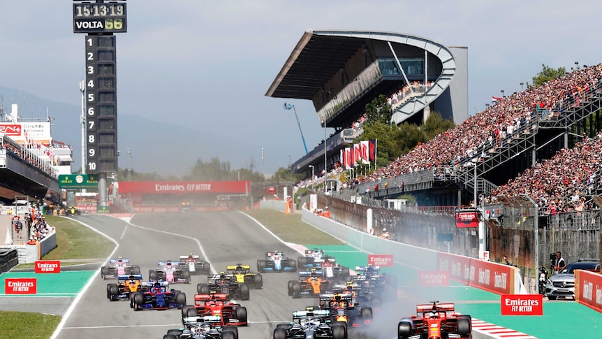 A group of Formula 1 cars race around a corner in front of large crowds.