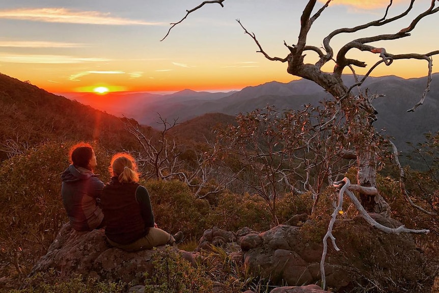 Two women sit on a rock on a mountain surrounded by bushland as the sun sets over mountains in the distance.