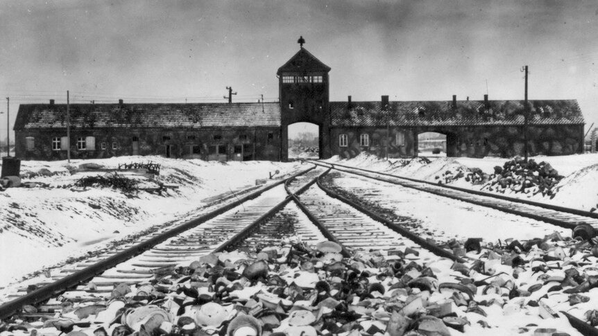 A grainy black-and-white photo of the entrance of Auschwitz concentration camp with snow on the tracks.